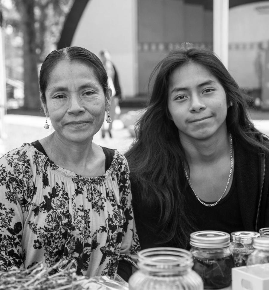 Two women smile at the camera behind a table with mason jars at an outdoor market or community event.