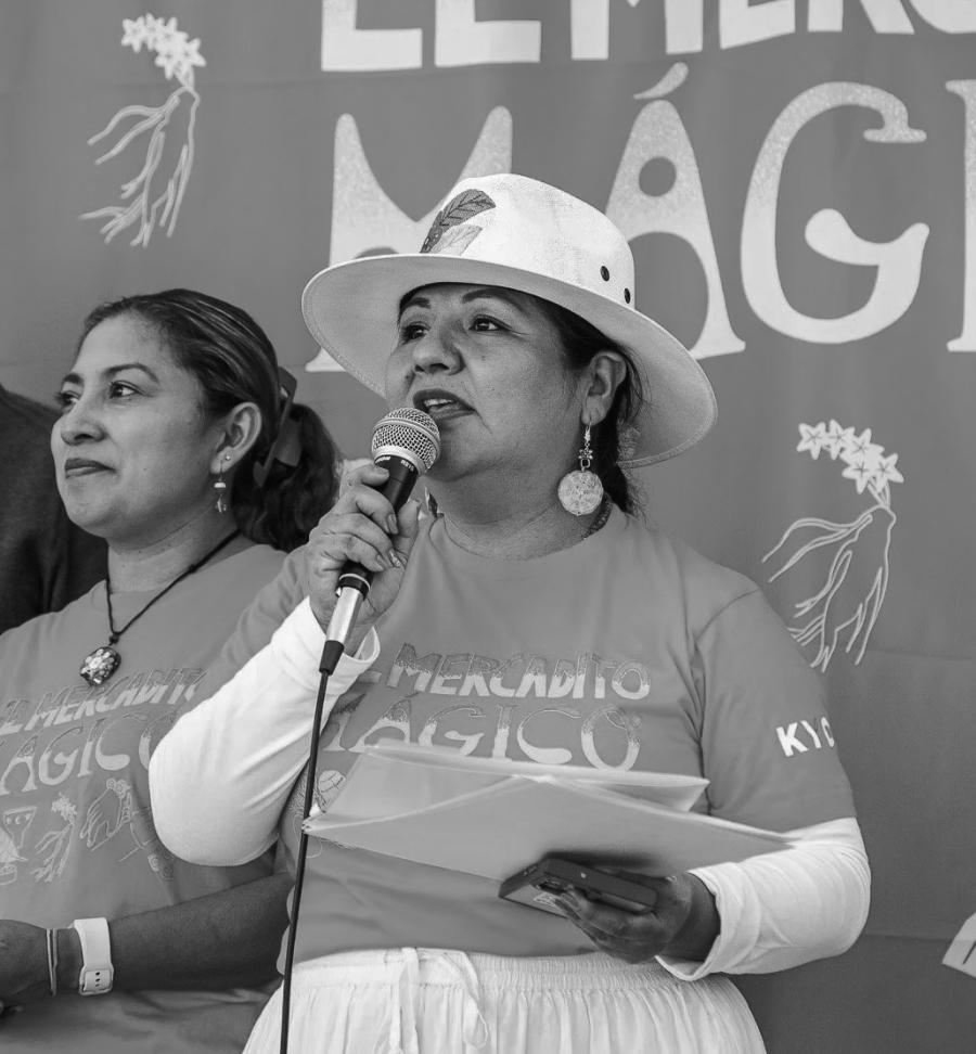 Deisy Gutierrez speaks into a microphone wearing a wide-brimmed hat and El Mercadito Mágico t-shirt while holding papers at an outdoor event with another woman in a matching shirt visible behind her.