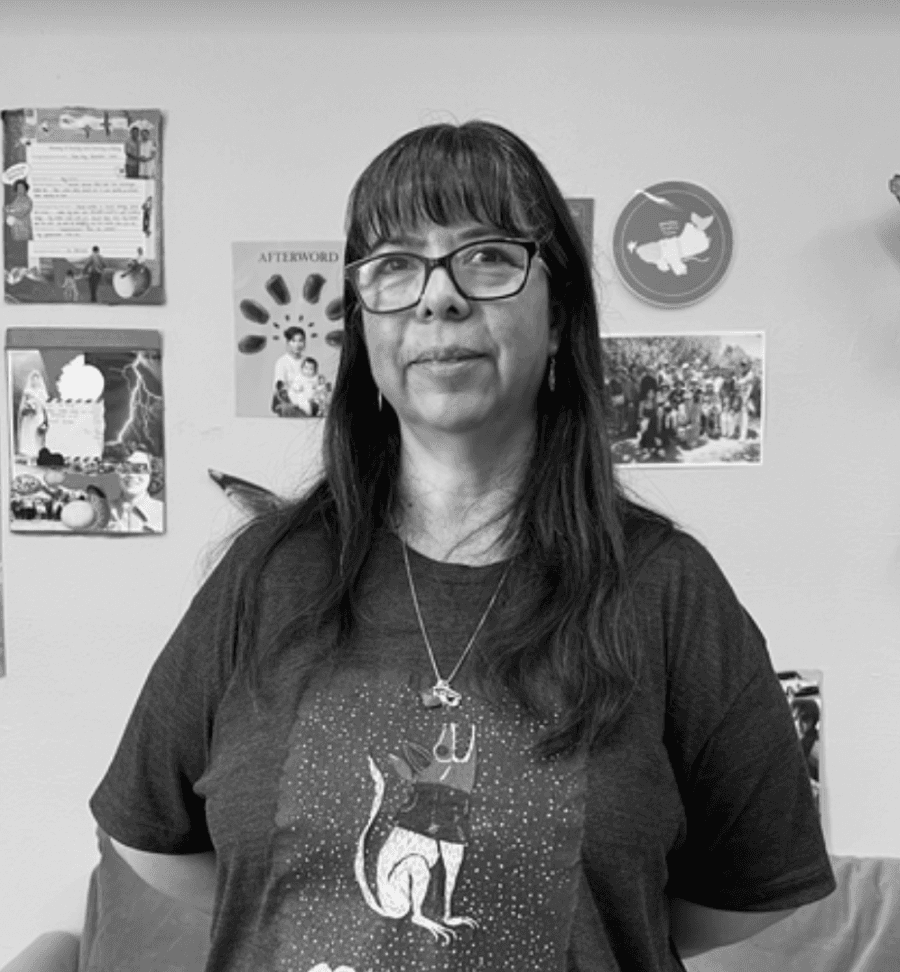 Edith Rincon wearing glasses and a graphic t-shirt stands in front of a wall covered with pinned photos and artwork.