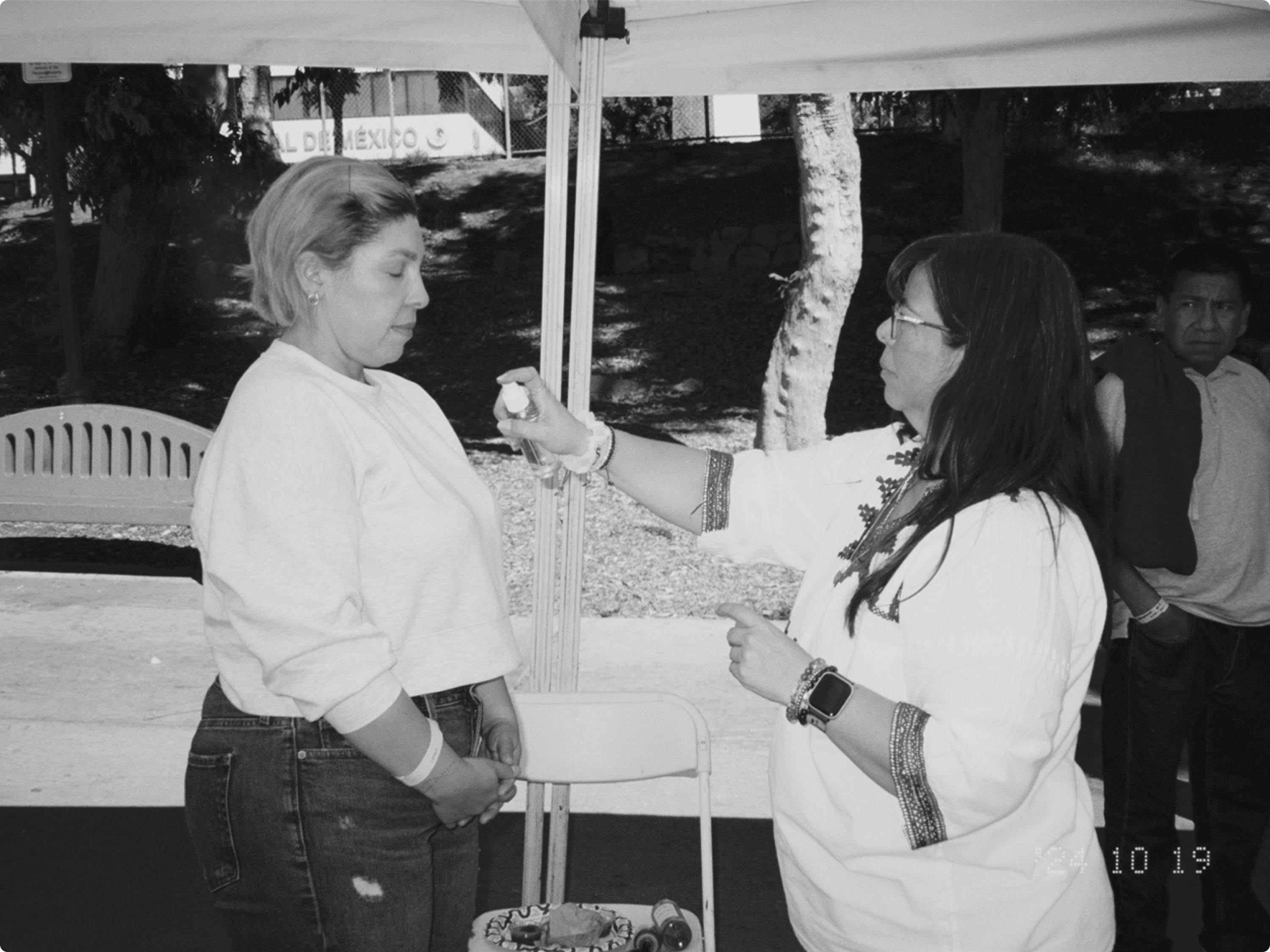 A woman in embroidered clothing applies a remedy to another woman's arm under an outdoor canopy tent.