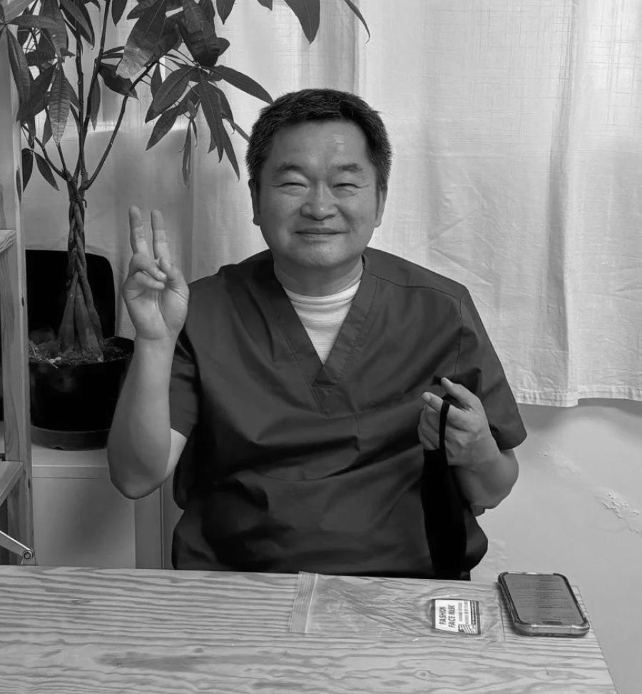 Han Jik Kim in dark medical scrubs sitting at a desk making a peace sign and smiling with a potted plant and white curtain in the background.