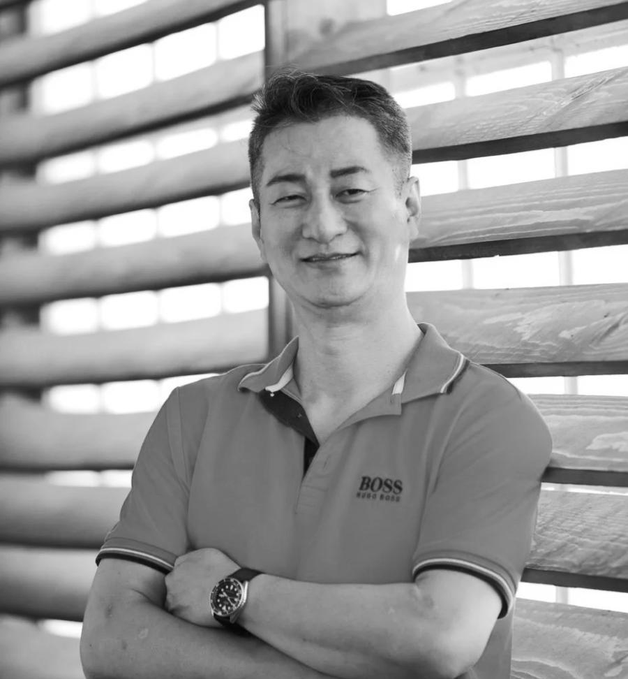 Headshot of Sae-Joon Oh smiling with arms crossed wearing a polo shirt in front of wooden louvered panels.