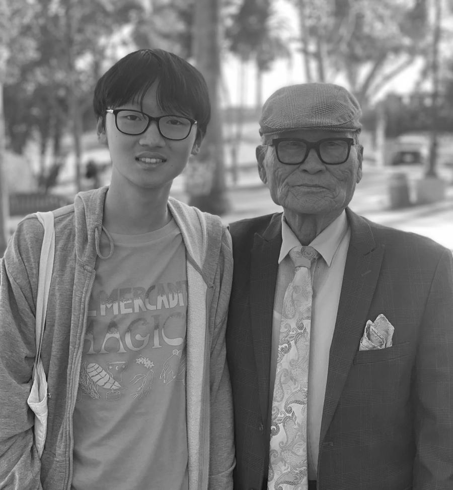 A young person wearing glasses and a hoodie stands beside an elderly man in a suit jacket, flat cap and patterned tie outdoors with palm trees in the background.